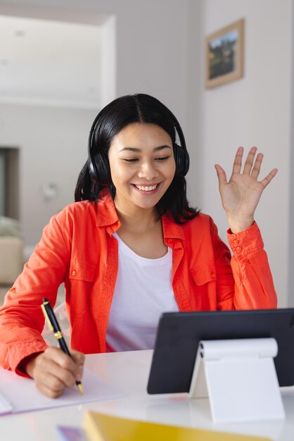 Vertical de una mujer birracial feliz con auriculares saludando, haciendo videollamadas con tableta en casa, copiando espacio. Trabajar desde casa, tecnología, comunicación y concepto de estilo de vida.