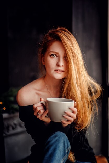 Vertical de una mujer joven adulta con pelo rojo en suéter negro sosteniendo una gran taza de bebida mirando a la cámara disfrutando de buenos días dentro de la casa con fondo gris oscuro borroso