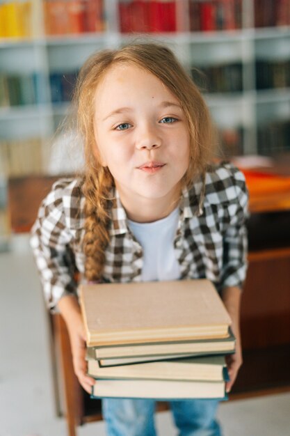 Vertical de la niña de la escuela del niño de primaria juguetona sosteniendo la pila de libros en la biblioteca en