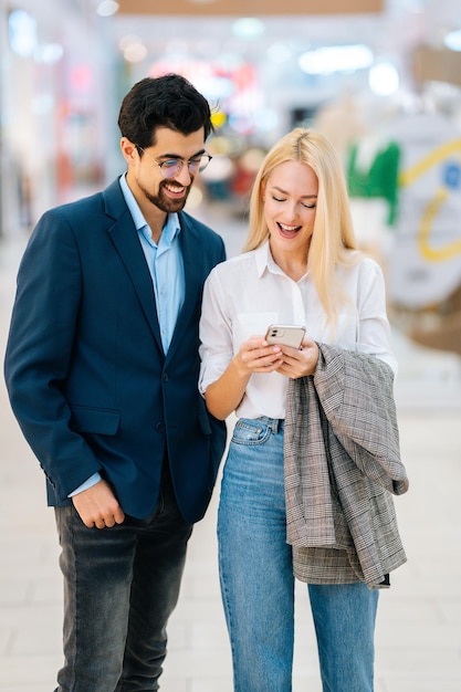 Vertical de pareja joven feliz enfocada mediante teléfono móvil de pie en el pasillo del centro comercial