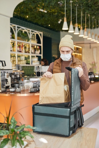 Vertical del repartidor con máscara mientras se empacan los pedidos de comida para llevar a la bolsa en el interior del café