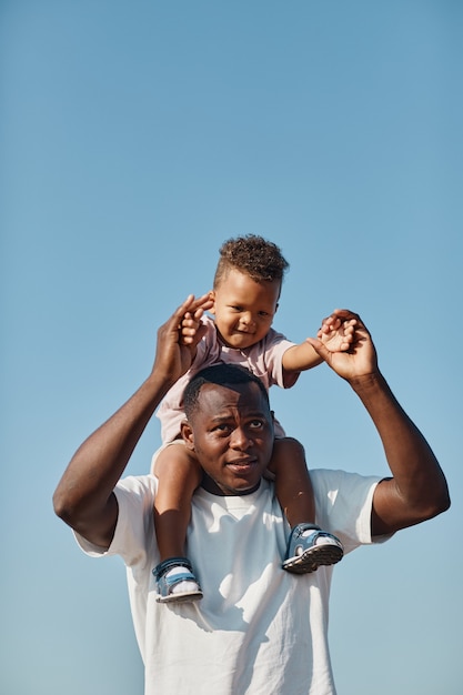 Vertical retrato de feliz padre afroamericano con hijo en hombros contra el cielo azul mientras e ...