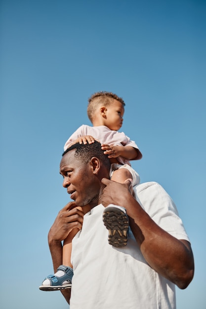 Vertical retrato de feliz padre afroamericano con hijo en hombros contra el cielo azul mientras e ...