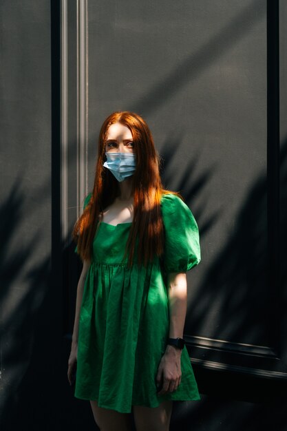 Foto vertical retrato de mujer joven atractiva seria con máscara médica de pie en las calles de la ciudad en un día soleado de verano, mirando a otro lado, listo para la campaña de vacunación, fondo oscuro.