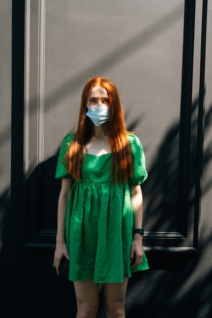 Vertical retrato de mujer joven atractiva seria vistiendo máscara médica de pie en las calles de la ciudad