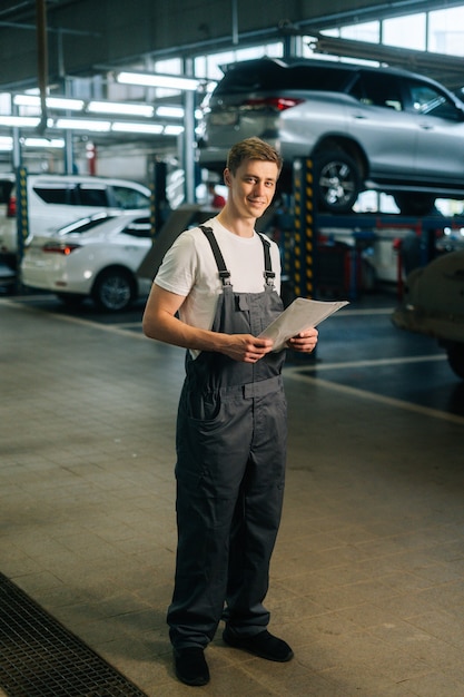 Vertical retrato de sonriente apuesto joven mecánico masculino vistiendo uniforme sosteniendo portapapeles de pie ...