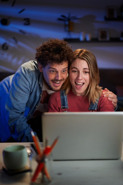 Vertical sorprendido pareja caucásica mirando feliz en la computadora sentado en el escritorio en la sala de estar oscura