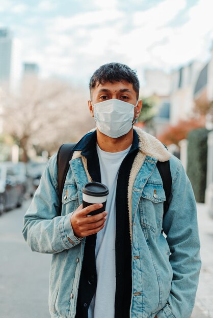 Vertikal eines jungen argentinischen stilvollen Mannes in einer Maske, die eine Tasse Kaffee auf der Straße hält