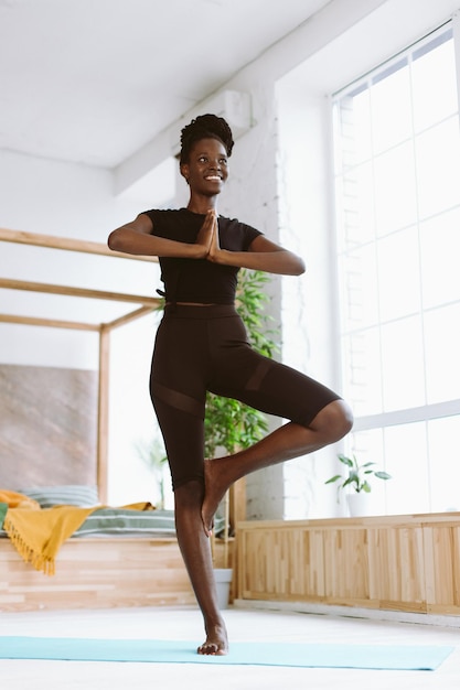 Vertikal lächelnde afroamerikanische Frau mit Dreadlocks, die Yoga-Baumhaltung Asana auf der Matte zu Hause praktiziert Niedrige Winkelansicht
