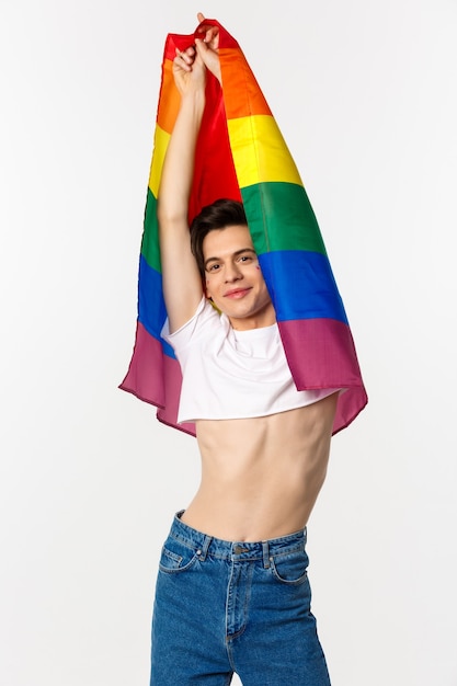 Vertikale Ansicht des schönen androgynen schwulen Mannes, der Regenbogenfahne erhebt und glücklich lächelt, im Erntedach und in den Jeans gegen Weiß steht.