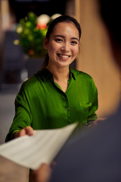 Vertikale Aufnahme einer jungen glücklichen asiatischen Büroangestellten, die ihr einige Dokumente gibt
