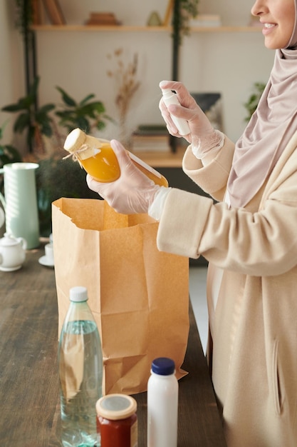 Vertikale Aufnahme einer modernen jungen muslimischen Frau, die Schutzhandschuhe trägt und die gelieferten Dinge t desinfiziert