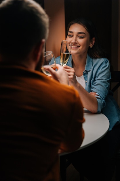 Vertikale Aufnahme eines glücklichen jungen Paares, das mit Champagner an einem Tisch mit Kerze in einem gemütlichen dunklen Raum klirrt. Liebender Mann und Frau feiern Jubiläum, Valentinstag, genießen romantisches Date