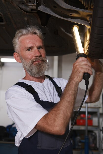 Vertikale Aufnahme eines leitenden Automechanikers, der eine Lampe hält und das Rad eines Autos auf dem Aufzug untersucht