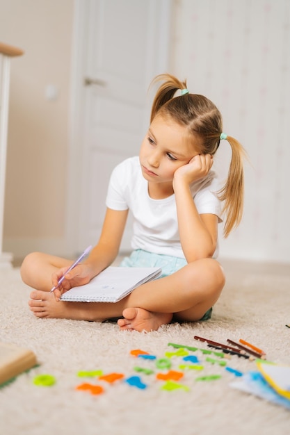 Vertikale Aufnahme eines müden kleinen Mädchens, das während des Homeschoolings zu Hause auf dem Boden sitzt und auf den Laptop-Bildschirm schaut