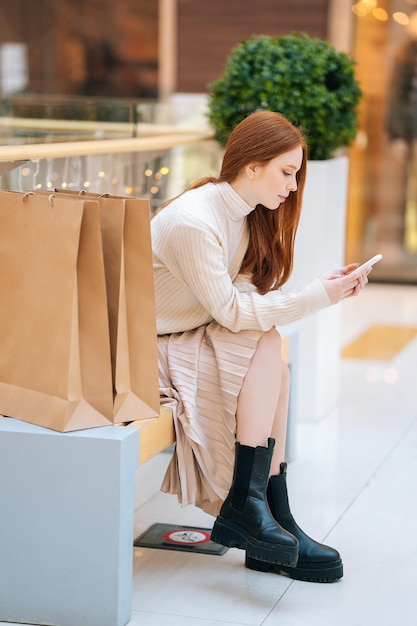 Vertikale Aufnahme in voller Länge einer fokussierten, süßen jungen Frau, die mit einem Mobiltelefon auf einer Bank mit Papier sitzt