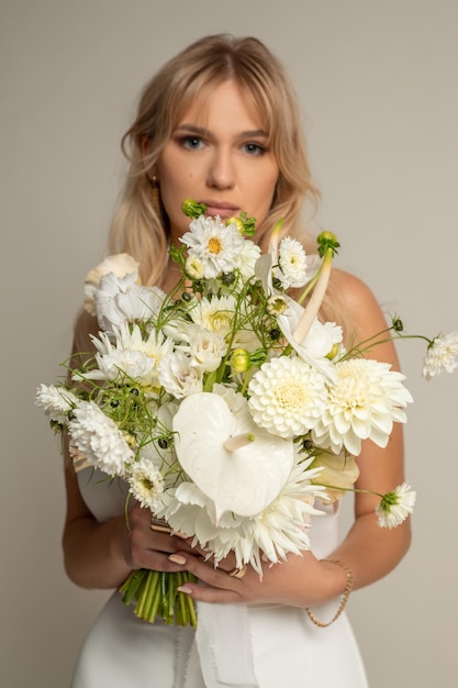 Vertikale, charmante, entspannte, blonde Frau, Braut in hellweißem Kleid, bedeckt mit einem großen weißen Blumenstrauß