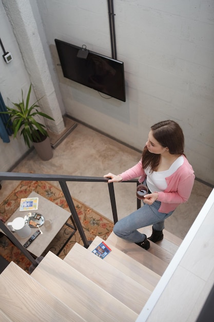 Vertikale Draufsichtaufnahme einer Frau, die die Treppe in ihrem Hotelzimmer mit einer Tasse Tee hinaufgeht
