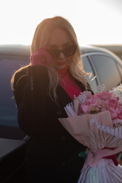 Vertikale Nahaufnahme, blonde Geschäftsfrau, die über einer Sonnenbrille schaut, die einen noblen rosa Rosenblumenstrauß gegen ein schwarzes Auto hält