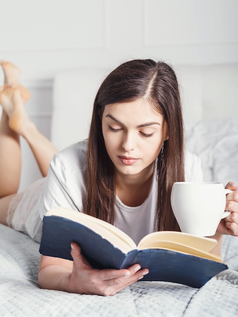 Vertikale Porträtansicht einer reizenden jungen Frau, die ein Buch liest und Kaffee auf dem Bett trinkt