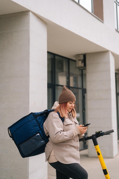 Vertikale Schnittaufnahme einer Kurierin mit großem Thermo-Rucksack mit Smartphone, die mit Elektroroller auf einer städtischen Straße steht