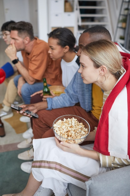 Vertikale Seitenansicht auf eine vielfältige Gruppe junger Leute, die zu Hause Sportspiele mit Popcorn und Snacks beobachten