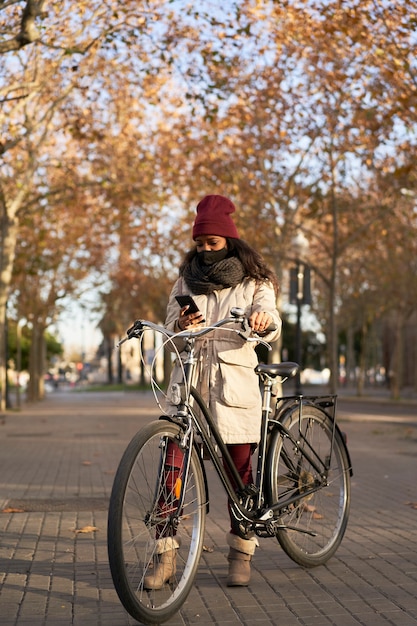 Vertikaler Schuss einer schwarzen Frau in der Winterkleidung, die mit einem Fahrrad steht, das am Telefon in einer Stadt bei Sonnenuntergang spricht.