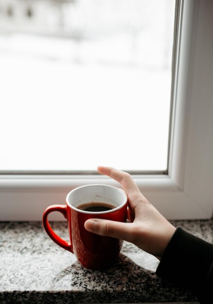 Vertikaler Schuss einer weiblichen Hand, die eine Tasse heißen Kaffee auf einem Fensterbrett hält