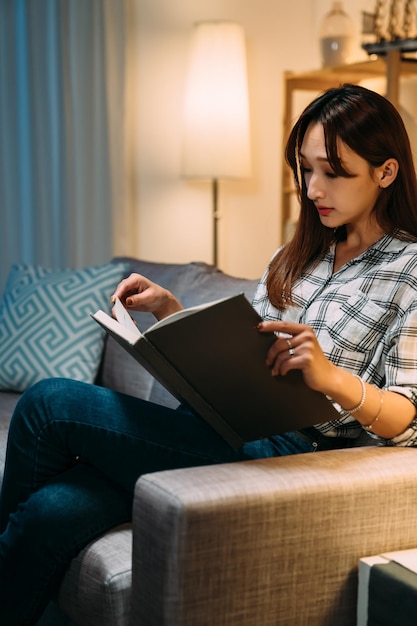 vertikaler schuss elegantes asiatisches mädchen genießt das lesen eines buches auf der couch in einem ruhigen wohnzimmer zu hause während der nachtzeit.