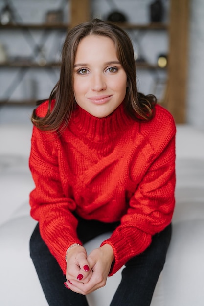 Vertikaler Schuss von angenehm aussehender Frau in lässigem rotem Strickpullover hat Maniküre genießt häusliche Atmosphäre hat Make-up-Posen im Innenbereich Schöne Frau mit dunklem Haar verbringt Freizeit zu Hause