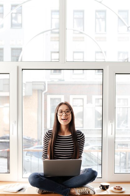 Vertikales Bild der asiatischen Frau auf Fensterbank mit Laptop