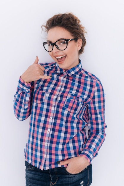 Vertikales Bild der Frau mit Brille und kariertem Hemd, das Telefonschild mit Arm in der Tasche zeigt. Isolierte graue Wand