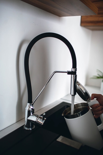 Vertikales Bild der Hand des Mannes, die einen Wasserkocher mit geöffneter Oberseite hält und etwas Wasser aus dem Wasserhahn eingießt. Brauchen Sie, um Wasser zu erhitzen. Moderne Küche mit guter Ausstattung und Sanitär