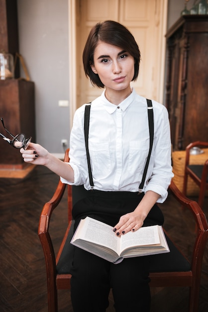 Vertikales Bild der hübschen Autorin, die mit Buch auf den Beinen auf einem Stuhl sitzt und eine Brille in der Hand hält