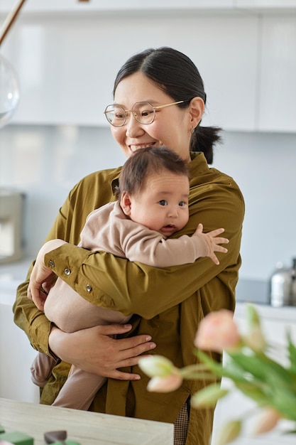 Vertikales Bild einer glücklichen asiatischen Mutter, die ihr Baby in den Armen hält und in der Küche steht