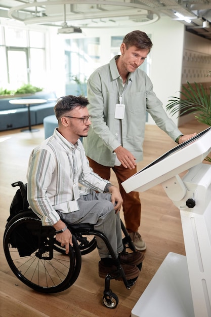 Vertikales Bild eines Mannes, der einem behinderten Mann im Büroraum beim Online-Eintrag hilft