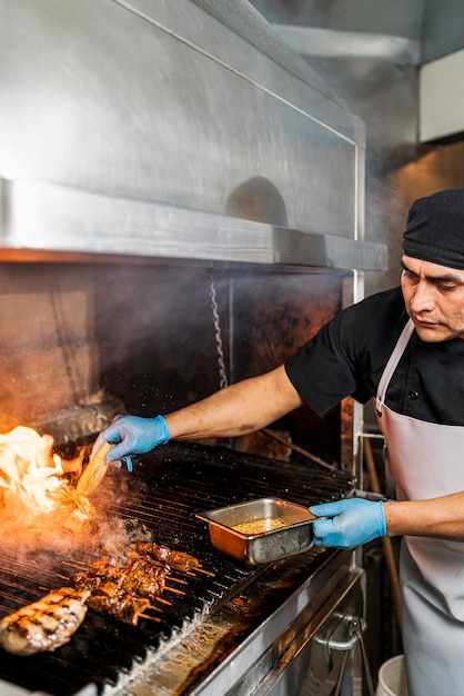 Vertikales Bild eines professionellen Kochs, der ein Stück Fleisch auf dem Grill mit Gewürzen bedeckt, wobei eine große Flamme aus dem Grill kommt.