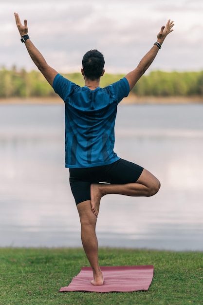 Vertikales Foto des Rückens eines Mannes, der auf einem Bein steht und eine Yoga-Pose mit Blick auf einen See in einem Park macht