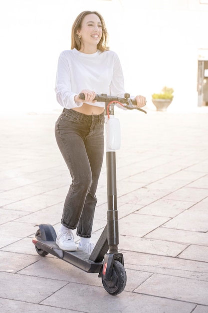 Vertikales Foto einer glücklich lächelnden positiven Frau, die an einem sonnigen Tag einen Tretroller fährt