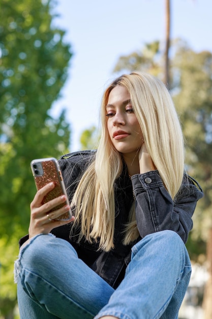 Vertikales Foto einer jungen Blondine, die im Park sitzt und auf ihr Telefon schaut