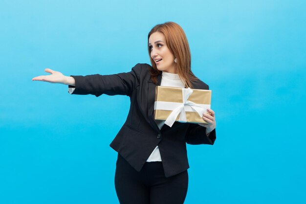 Vertikales Foto einer jungen Geschäftsfrau, die eine Geschenkbox hält und ihre Hand zur Seite zeigt