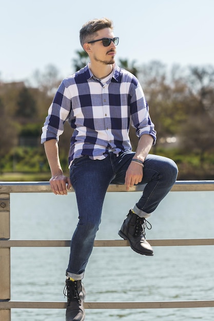 Vertikales Foto eines jungen kaukasischen Mannes, der auf dem Geländer eines schwimmenden Stegs neben einem Fluss sitzt