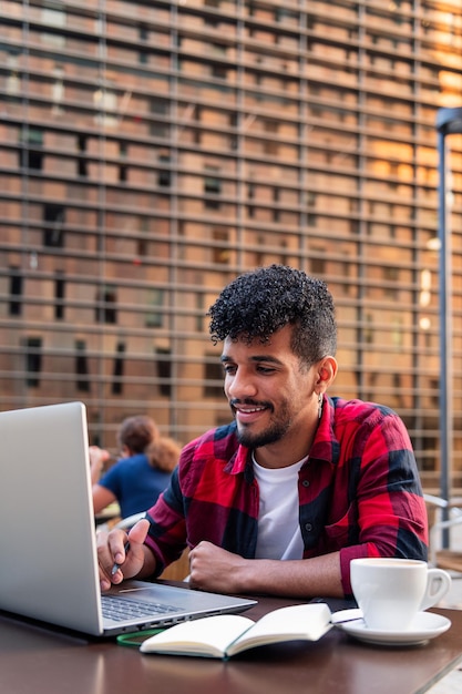 Vertikales Foto eines jungen lateinamerikanischen Studenten, der mit seinem Computer auf der Terrasse eines Cafés in der Stadt arbeitet, Konzept von Technologie und urbanem Lebensstil