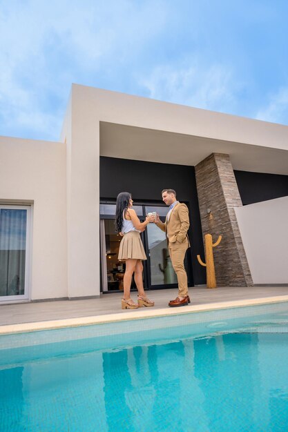 Vertikales Foto eines Paares, das in der Terrasse ihres neuen Luxushauses neben dem Pool toastet