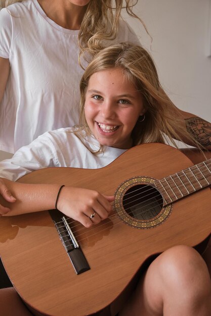 Vertikales Foto eines Porträts eines blonden Mädchens, das Gitarre spielt, während es vor der Kamera lächelt, mit einer Frau, die mit ihr auf einer Couch in einem Raum sitzt