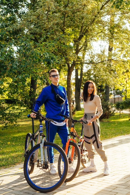 Vertikales Foto eines stilvoll lächelnden positiven Paares, das mit Sportfahrrädern im Park steht