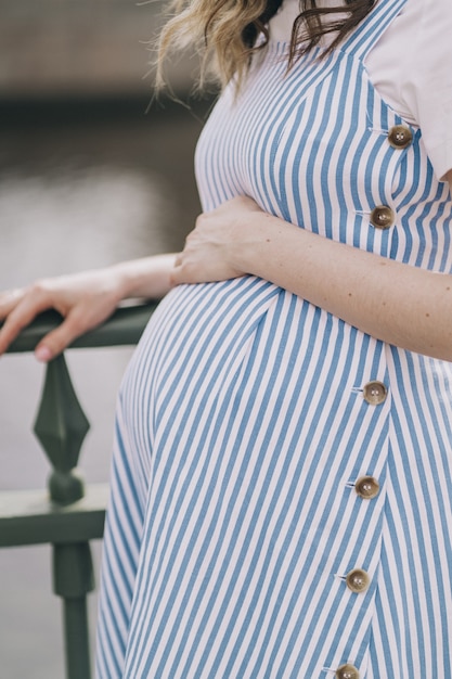 Foto vertikales foto mit einem schwangeren mädchen. nahaufnahme des bauches eines schwangeren mädchens. eine junge mutter streichelt ihren schwangeren bauch. schwangere frau in einem zarten blauen kleid. foto in hoher qualität