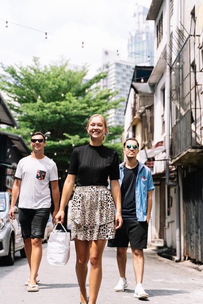 Vertikales Foto von drei jungen Menschen unterschiedlicher Ethnie und Geschlecht, die lächeln und eine Straße mit Autos, Bäumen und Lichtern entlanggehen, die hängen, während die Frau eine Tasche hält holding