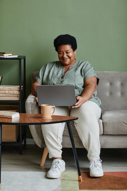 Vertikales Ganzkörperporträt einer schwarzen Seniorin, die zu Hause einen Laptop benutzt, während sie sich auf der Couch entspannt