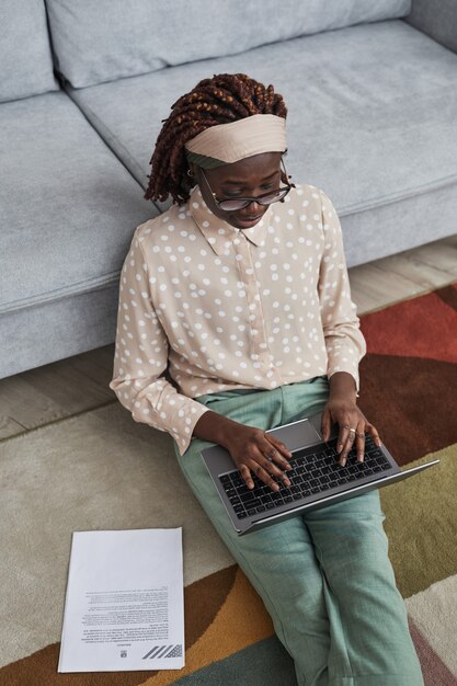 Vertikales Hochwinkelporträt einer modernen afroamerikanischen Frau, die von zu Hause aus arbeitet, während sie auf einem grafischen Teppich auf dem Boden sitzt und Laptop verwendet, Platz kopieren
